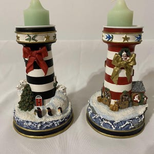 May include: Two Christmas-themed candle holders shaped like lighthouses. One is red and white with gold accents and the other is black and white. Both have a snowy base with penguins and polar bears.