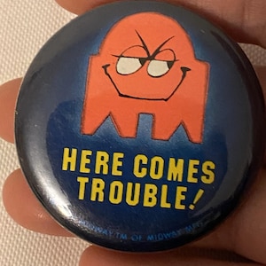 May include: A blue and orange button with a cartoon ghost and the text "Here Comes Trouble!"  A trademark symbol and the text "TM of Midway Mfg." are visible at the bottom of the button.