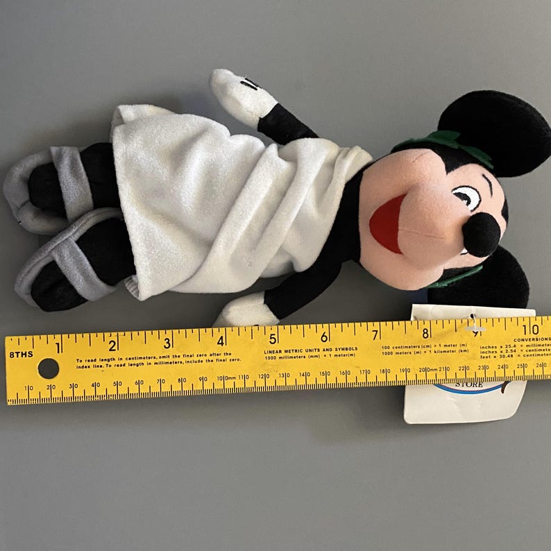 May include: A plush Mickey Mouse toy wearing a white toga and sandals. The toy is lying on its back with its arms outstretched. The toy is approximately 10 inches long.