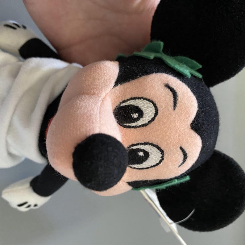 May include: A plush Mickey Mouse toy wearing a white toga and a green laurel wreath. The toy has black ears, a black nose, and embroidered eyes.