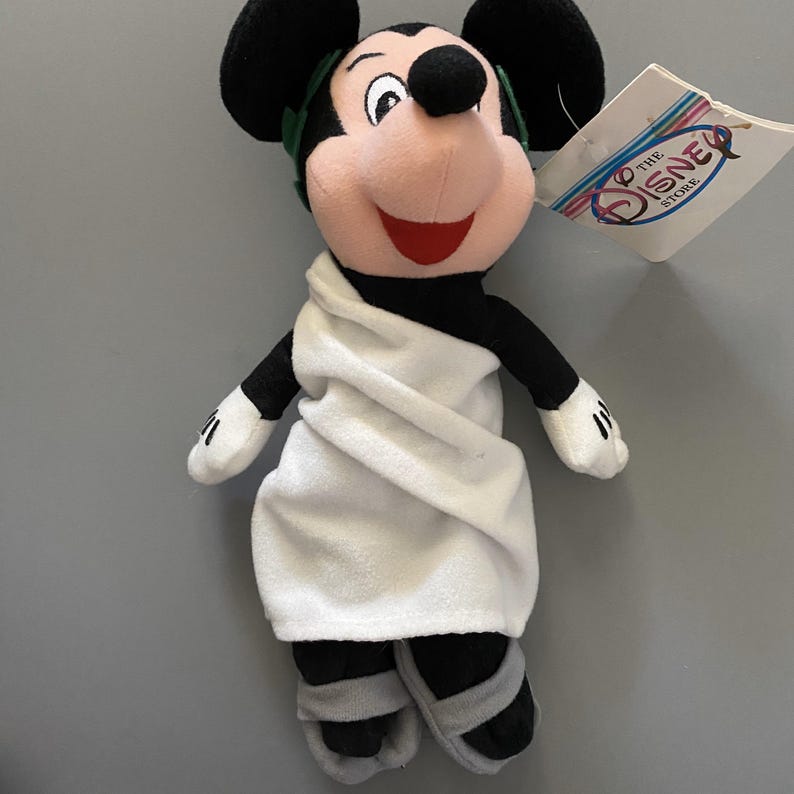 May include: A plush Mickey Mouse toy wearing a white toga and sandals. The toy has a green laurel wreath on its head and is holding a tag that says "The Disney Store".