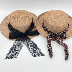 May include: Two straw sun hats with wide brims. One hat features a black lace bow and streamers, while the other has a leopard print scarf tied around the crown. The hats are tan and have a woven texture.