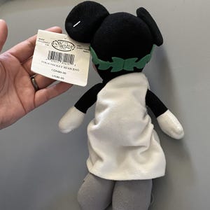 May include: A black and white plush Mickey Mouse wearing a white toga and a green laurel wreath. The plush toy is a bean bag and has a tag that reads "TOGA MICKEY BEAN BAG CDN$9.00 US$6.00".