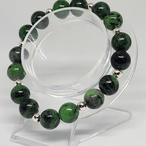 May include: A green and black gemstone bracelet with silver beads. The bracelet is made of round beads and is displayed on a clear plastic stand.