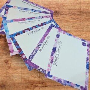 Może przedstawiać: Stos spersonalizowanych papierów listowych z marmurkowym wzorem w odcieniach fioletu, niebieskiego i bieli. Każda kartka ma wydrukowane imię i nazwisko "Meredith K. Herman, DO" czarnym pismem. Papier listowy ułożony jest na drewnianej powierzchni.