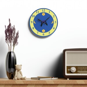 Puede incluir: Un reloj azul y amarillo con un dise&ntilde;o de luna y sol. La esfera del reloj tiene n&uacute;meros amarillos y una aguja negra. El reloj est&aacute; colgado en una pared blanca.