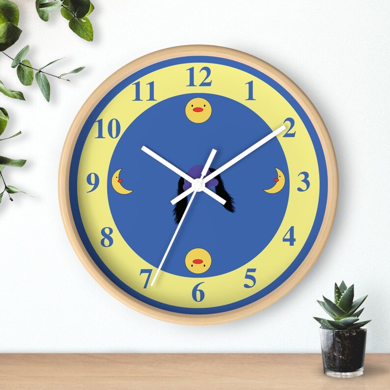 Puede incluir: Un reloj redondo de madera con una esfera azul y n&uacute;meros amarillos. El reloj tiene una aguja blanca y una silueta morada de una persona con un sombrero amarillo. La esfera del reloj tambi&eacute;n tiene lunas y patos amarillos.