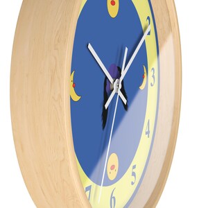 Puede incluir: Un reloj con marco de madera, con una esfera azul y n&uacute;meros amarillos. El reloj tiene una aguja de minutos blanca y una aguja de horas negra. Hay tres lunas amarillas con caras rojas en la esfera del reloj.