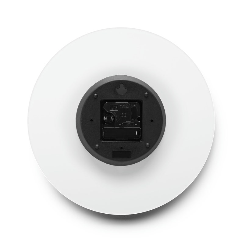 Puede incluir: La parte posterior de un reloj redondo blanco con un mecanismo de reloj negro. El mecanismo del reloj tiene un peque&ntilde;o gancho negro en la parte superior y un peque&ntilde;o bot&oacute;n negro en la parte inferior.