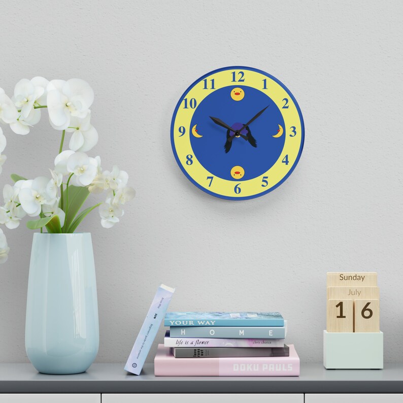 Puede incluir: Un reloj de pared azul y amarillo con un dise&ntilde;o de b&uacute;ho negro y patos amarillos en los n&uacute;meros 12 y 6. Las manecillas del reloj apuntan a las 10:30. El reloj est&aacute; colgado en una pared blanca. Una pila de libros y un calendario de madera con la fecha 16 de julio est&aacute;n en un estante debajo del reloj.