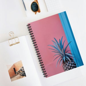 May include: Spiral-bound notebook with a pink cover featuring a blue pineapple illustration. The notebook is open, revealing blank pages. Gold-colored binder clip and sunglasses are also visible on a white surface.