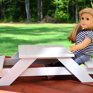 May include: A white wooden picnic table with a doll sitting on one of the benches. The table is on a red wooden surface.