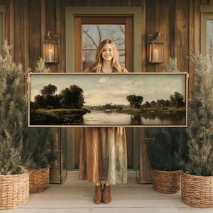 Op de afbeelding: Een ingelijst landschapsschilderij met een rivierlandschap met bomen en een bewolkte lucht. Het kunstwerk wordt gepresenteerd tegen een houten achtergrond, met decoratieve lantaarns en potplanten. Het schilderij is rechthoekig en heeft een lichtgekleurde lijst.