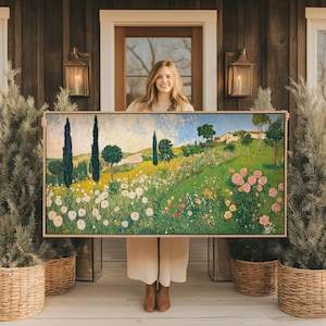 May include: A framed landscape painting featuring a vibrant field of flowers, rolling green hills, and tall cypress trees under a blue sky. The artwork has a light-colored wooden frame and depicts a sunny, pastoral scene.