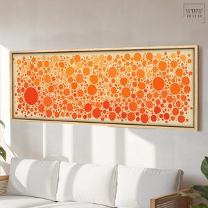 May include: Abstract art print featuring a pattern of orange and red circles of varying sizes against a cream background. The artwork is framed in a light wood frame and hangs above a white sofa.