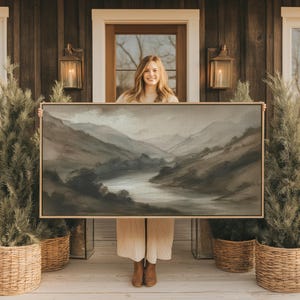 May include: A framed landscape painting depicting a river winding through a mountain range. The artwork features a muted color palette of grays and browns, with a gold-colored frame. The painting is held up in front of a wooden door.