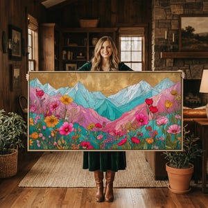 May include: A framed landscape art print featuring a colorful mountain scene with pink, blue, and gold hues. The artwork showcases a vibrant meadow of flowers in shades of pink, yellow, and red. The print is held up in front of a wooden interior.