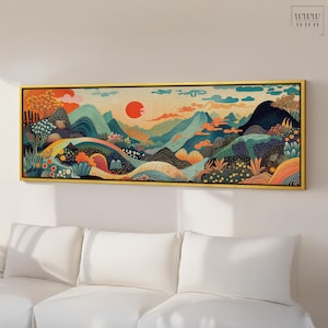 Op de afbeelding: Een ingelijst landschapsschilderij met een gestileerde bergscène met een rode zon en een palet van blauw, groen en oranje. Het kunstwerk wordt in een gouden lijst boven een witte bank getoond.