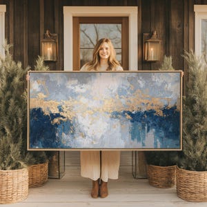Op de afbeelding: Abstract schilderij met een goud- en blauw kleurenpalet. Het kunstwerk heeft een mix van blauw, wit en goud, waardoor een getextureerd, gelaagd effect ontstaat. Het schilderij is ingelijst in een lichtgekleurde houten lijst, geschikt voor interieurdecoratie.