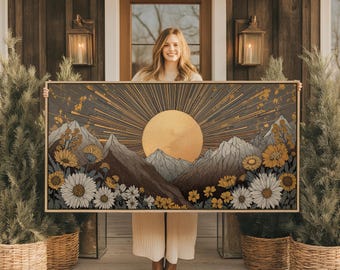 May include: A framed art print featuring a stylized mountain landscape with a large golden sunburst. The artwork includes mountains, flowers, and a color palette of gold, gray, and brown. The frame is a light wood color.