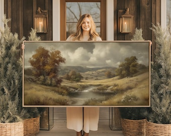 May include: A landscape painting in a gold frame depicts a serene river scene with rolling hills and trees. The artwork features a muted color palette of greens, browns, and grays, creating a vintage aesthetic. The painting is held up in front of a wooden door.