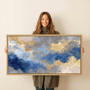 Peut inclure: Impression d'art abstrait dans un cadre en bois rectangulaire. L'œuvre présente un mélange de teintes bleues, dorées, grises et blanches, créant un effet nuageux ou atmosphérique. La peinture est tenue par une personne.