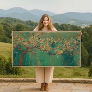 Può includere: Una stampa artistica incorniciata raffigurante un albero stilizzato con un tronco e rami rossi, adornato con forme circolari arancioni e gialle su uno sfondo verde e turchese. L'opera d'arte è tenuta davanti a una persona.