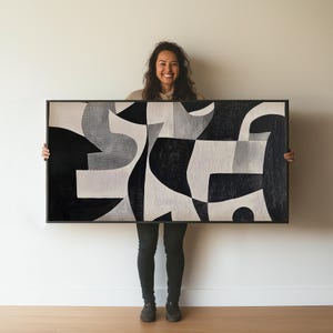 以下が含まれることがあります： 黒、白、グレーの幾何学的な形をした大きな抽象画。絵は黒い額縁に入っていて、人が持っています。