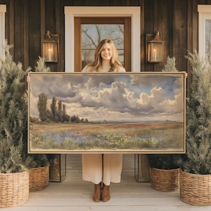May include: A large rectangular landscape painting in a thin wooden frame, depicting a vast field of wildflowers under a dramatic sky filled with heavy, textured clouds, with tall dark trees on the left and a distant horizon line.