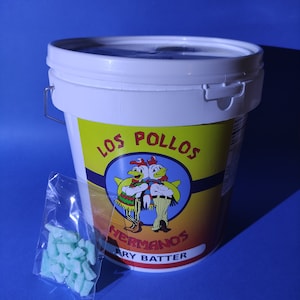 Peut inclure: Un seau en plastique blanc avec une étiquette jaune et rouge qui dit "Los Pollos Hermanos Fry Batter". L'étiquette présente une image de dessin animé de deux poulets portant des sombreros. Un petit sac en plastique contenant un mélange de pâte à frire verte est à côté du seau.