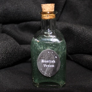 Puede incluir: Una pequeña botella de vidrio con un tapón de corcho llena de un líquido verde. La botella tiene una etiqueta que dice "Basilisk Venom".