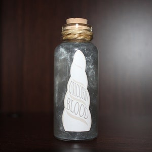 Op de afbeelding: Een klein glazen flesje met een kurkstop en een wit etiket met de tekst "Unicorn Blood". Het flesje is gevuld met een glinsterende, zilveren vloeistof.