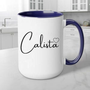 May include: White ceramic mug with a blue rim and handle. The mug has a black cursive design that says "Calista" with a small heart next to it.