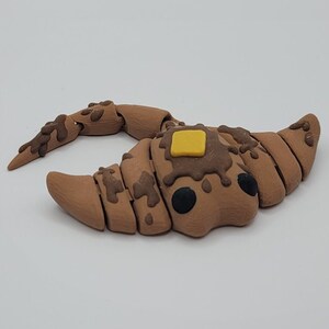 May include: A brown, segmented croissant-shaped sculpture with dark brown drizzles and a yellow square of butter on top. The sculpture has two black circular eyes and is set against a white background.