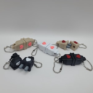 May include: Five different keychains with a heart design. The keychains are made of plastic and come in a variety of colors, including black, white, and beige. Each keychain has a small heart-shaped charm attached to it.