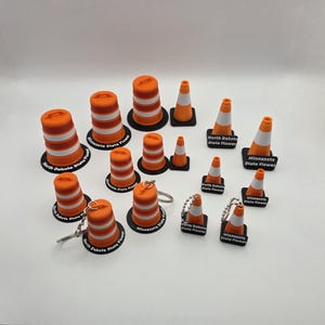 May include: A collection of miniature orange and white traffic cones and barrels. Some have keychains attached. The bases are black and have text that reads "North Dakota State Flower" and "Minnesota State Flower".