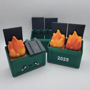 May include: Two green toy dumpsters with black lids. One dumpster has a smiling face and a fire inside. The other dumpster has the year 2025 printed on the front and a fire inside.
