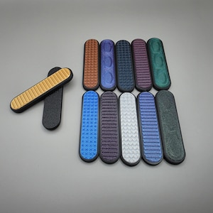 May include: An assortment of colorful, textured, oval-shaped objects. The objects are in various colors, including gold, black, blue, purple, and green. The textures vary, with some having a grid pattern and others a series of raised dots. The objects are approximately 4 inches long.
