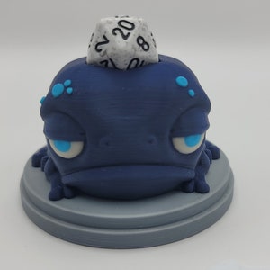 May include: A blue 3D printed frog figurine with a white 20-sided die on its head. The frog has blue eyes and a grumpy expression.