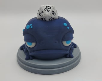 May include: A blue 3D printed frog figurine with a white 20-sided die on its head. The frog has blue eyes and a grumpy expression.