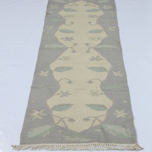 May include: A rectangular, hand-woven rug with a central cream-colored design framed by a gray border. The rug features floral and leaf patterns in light blue and cream, with fringed edges. The rug is approximately 60 inches long.