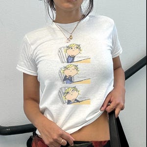 Peut inclure: T-shirt blanc avec une illustration de dessin animé d'une fille à un bureau en trois panneaux. La fille a les cheveux blonds et porte une robe bleue. Le t-shirt à manches courtes a un col rond.