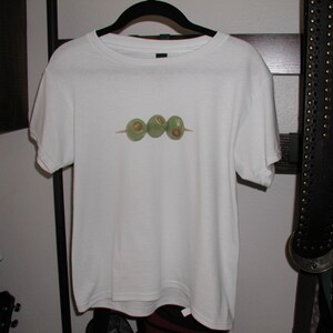K&ouml;nnte beinhalten: Wei&szlig;es T-Shirt mit einem Aufdruck von drei gr&uuml;nen Oliven auf einem Spie&szlig;.