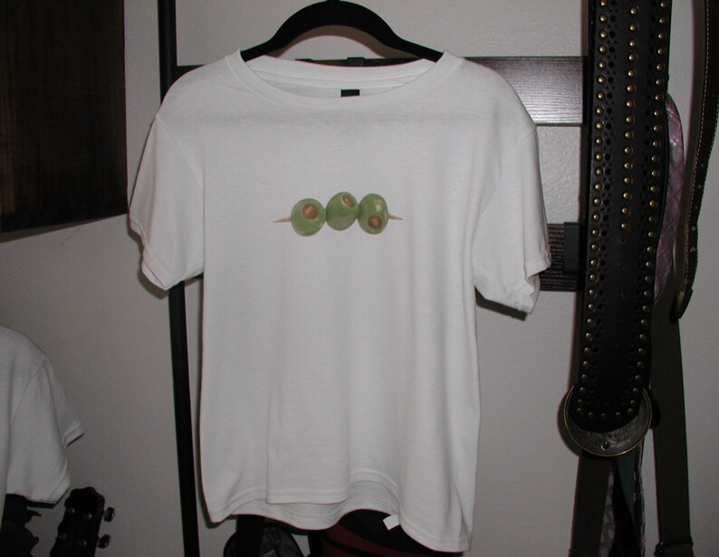 K&ouml;nnte beinhalten: Wei&szlig;es T-Shirt mit einem Aufdruck von drei gr&uuml;nen Oliven auf einem Spie&szlig;.