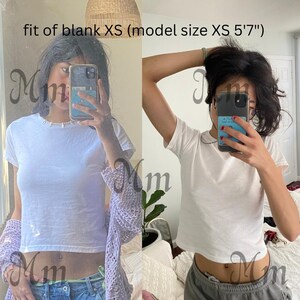 May include: Two images of a person wearing a white, short-sleeved t-shirt. The text "fit of blank XS (model size XS 5'7")" is visible. The person is holding a phone in front of their face.