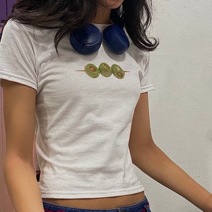 K&ouml;nnte beinhalten: Wei&szlig;es, bauchfreies T-Shirt mit einem Aufdruck von drei gr&uuml;nen Oliven auf einem Spie&szlig;.