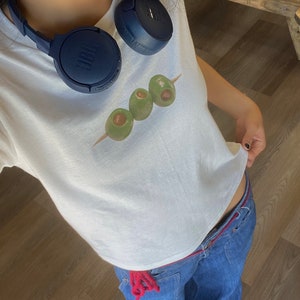 K&ouml;nnte beinhalten: Ein wei&szlig;es T-Shirt mit einem Aufdruck von drei gr&uuml;nen Oliven auf einem Spie&szlig;. Das Shirt wird mit einer blauen Jeans und einem blauen Kopfh&ouml;rer um den Hals getragen.