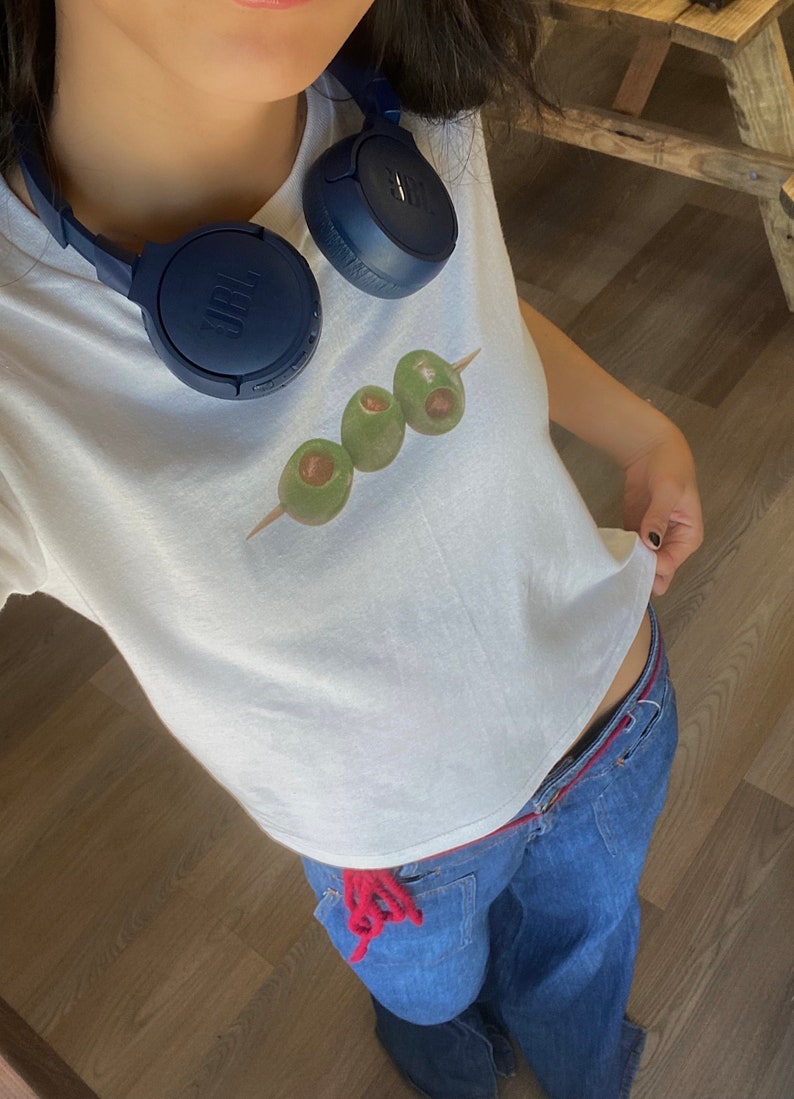 K&ouml;nnte beinhalten: Ein wei&szlig;es T-Shirt mit einem Aufdruck von drei gr&uuml;nen Oliven auf einem Spie&szlig;. Das Shirt wird mit einer blauen Jeans und einem blauen Kopfh&ouml;rer um den Hals getragen.