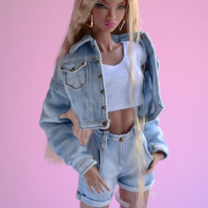 May include: A doll with long blonde hair wearing a light blue denim jacket and matching denim shorts. The doll has brown eyes and is wearing a white vest top.