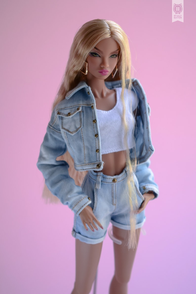 May include: A doll with long blonde hair wearing a light blue denim jacket and matching denim shorts. The doll has brown eyes and is wearing a white vest top.